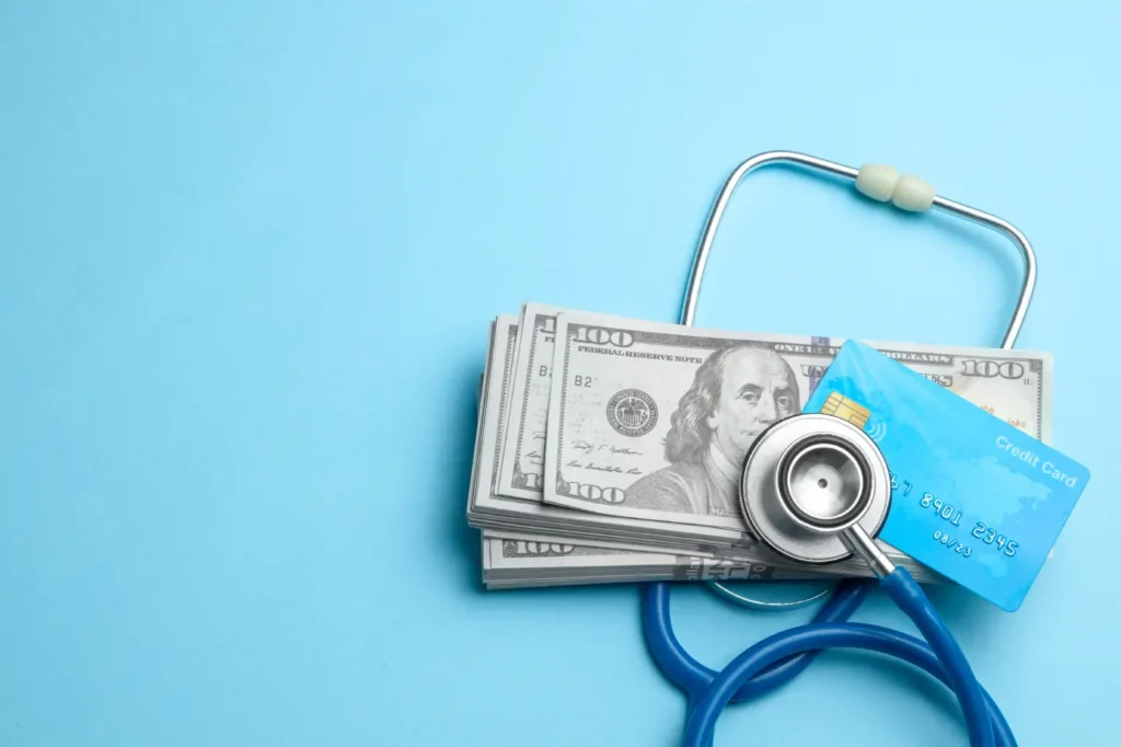A stethoscope and credit card resting on a stack of hundred dollar bills, representing the hidden costs of choosing health insurance based on price alone.