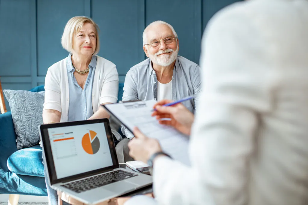 Understanding asrs: A smiling senior couple meets with a financial advisor who is reviewing documents on a clipboard, with a laptop displaying a pie chart visible in the foreground.